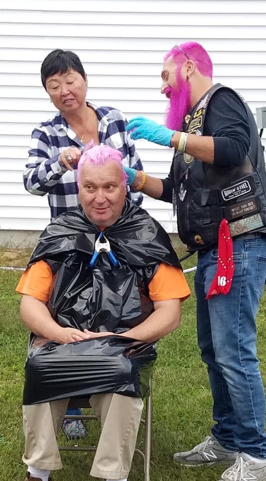 A sponsor having his hair dyed bright pink outdoors as two people help